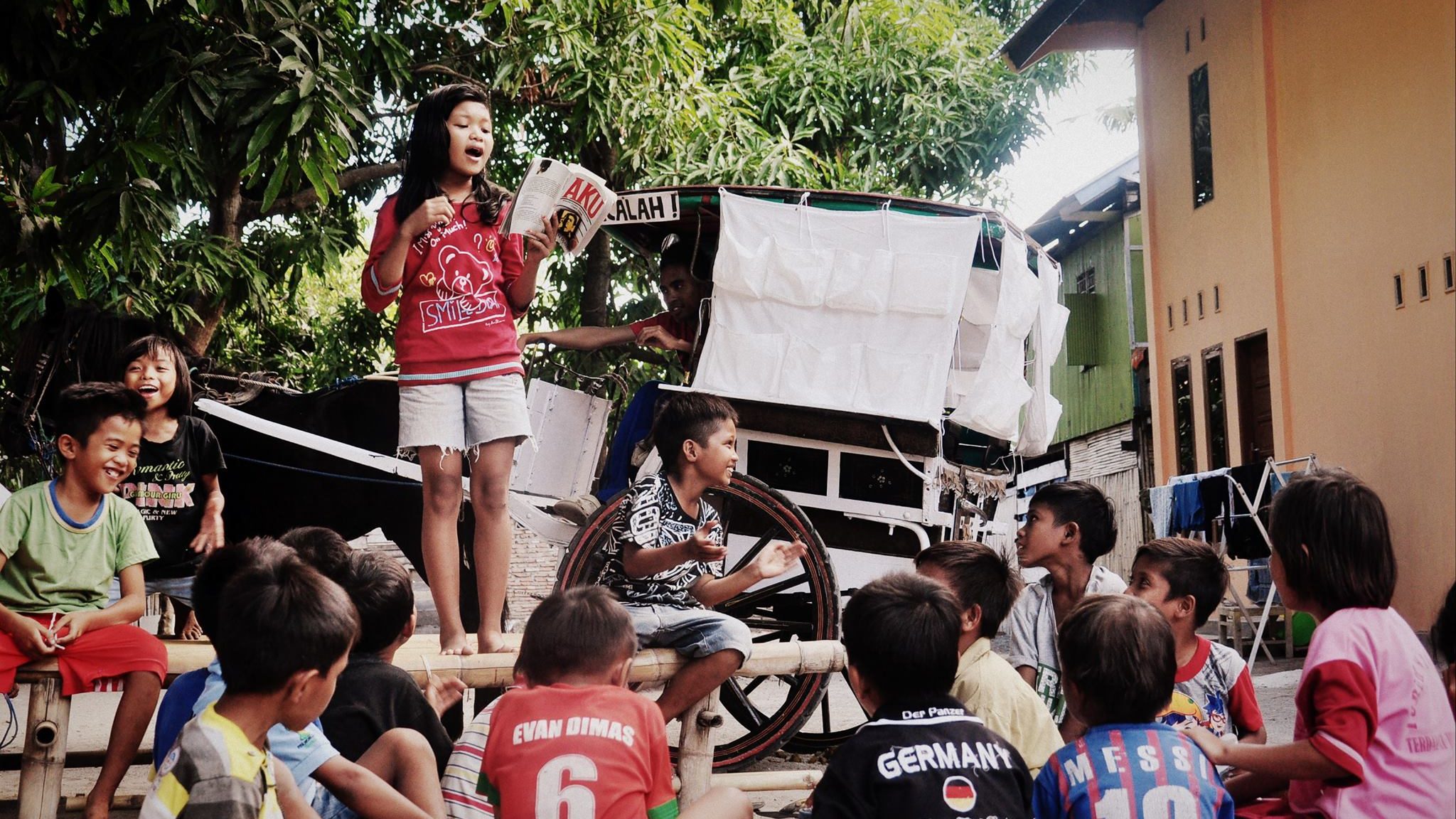 A girl is standing on a platform in front of a horse-cart and some trees, she is surrounded by other children sitting, laughing and listening as she reads out-loud from a book.